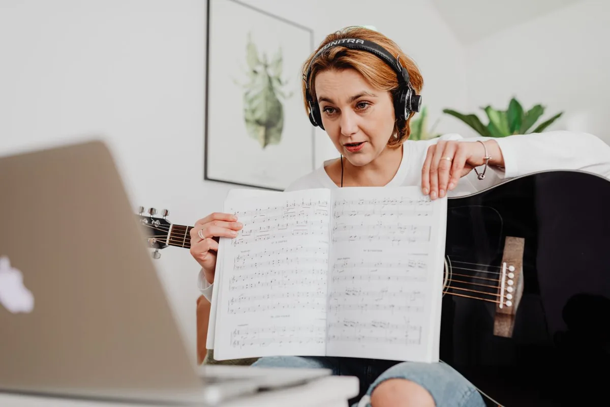 Mulher com guitarra e auscultadores a mostrar uma partitura junto de um computador