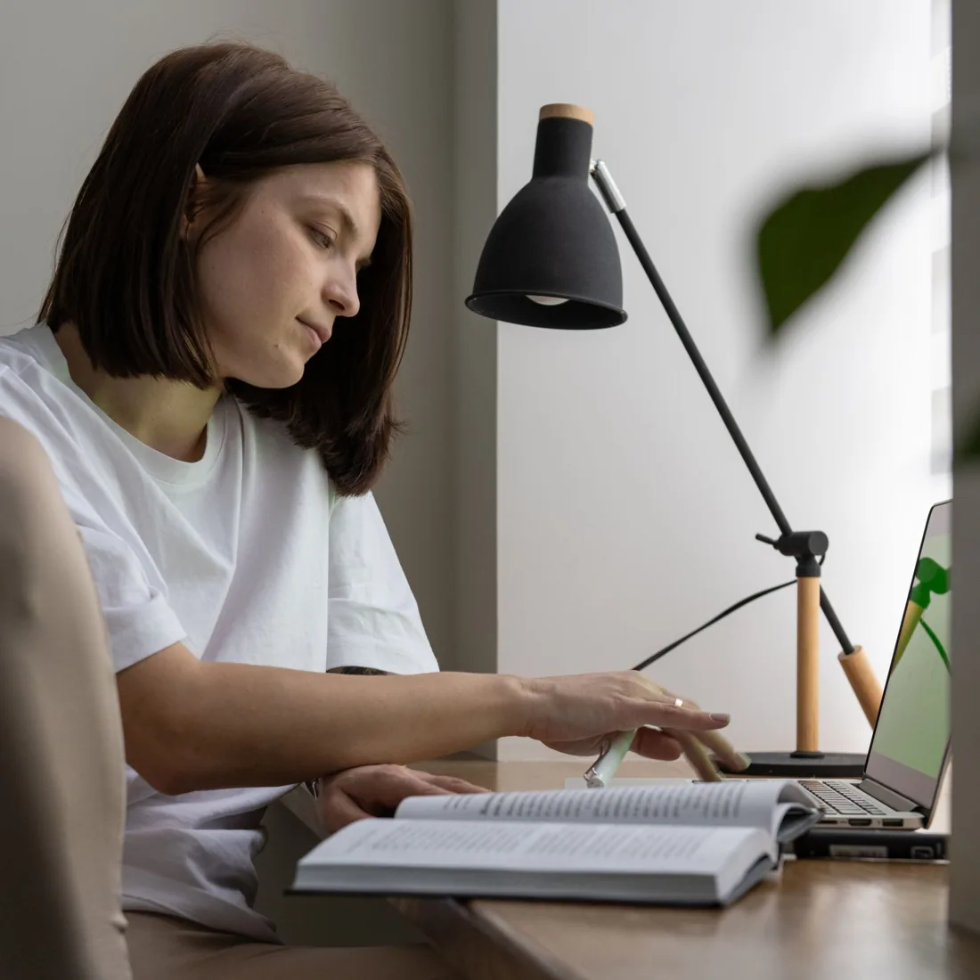 Mulher a consultar um livro aberto enquanto trabalha num computador portátil